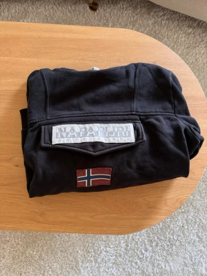 Svart hoodie från Napapijri - Säljer en svart hoodie från Napapijri med stor logga och norsk flagga på bröstet. Storlek M