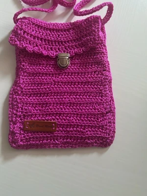 Rosa virkad axelväska med lock - Snygg axelväska i starkt rosa färg, virkad i garn med ett dekorativt lock och metallspänne framtill. Väskan har en lång axelrem och en liten läderdetalj med text på framsidan. Perfekt för dig som vill ha en unik och handgjord accessoar.