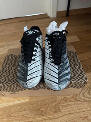 Svarta och vita fotbollsskor från Umbro - Snygga fotbollsskor från Umbro i svart och vitt med grafiskt mönster och vita detaljer. Skorna har snörning och låg profil, perfekt för dig som vill ha stilrena och funktionella skor på planen. Tillverkade i syntetmaterial för bra grepp och kontroll.