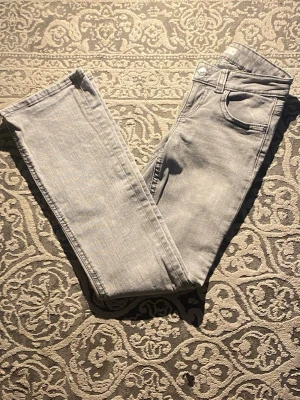Ljusgrå raka jeans i denim - Säljer ett par ljusgrå jeans med raka ben och klassisk femficksmodell. Jeansen har en unik bakficka med lock och knapp, vilket ger en extra detalj. Tillverkade i mjukt denimtyg, perfekta för en clean och stilren look.