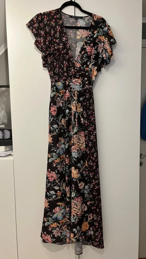 Svart långklänning med blommigt mönster - Långklänning med svart botten och färgglatt blommigt mönster i rosa, blått, gult och grönt. Klänningen har v-ringning, volangdetaljer vid ärmarna och ett omlottliv. Perfekt för dig som vill ha en feminin och flowy look.