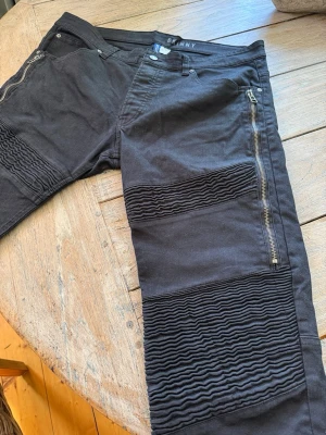 Svarta skinny jeans med dragkedjor - Säljer ett par svarta skinny jeans med coola detaljer som dragkedjor på sidorna och ribbade partier över knäna. Jeansen har fem fickor och är tillverkade i stretchigt material för en tajt passform. Storlek 36, 180/90A