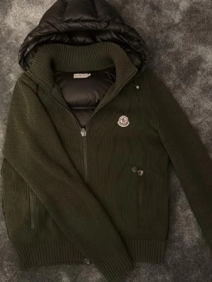 Grön stickad jacka från Moncler - Mörkgrön stickad jacka från Moncler med quiltad huva och dragkedja framtill. Jackan har ribbade muddar, två fickor med dragkedja och Moncler-logga på bröstet. Perfekt för kyliga dagar och ger en snygg, avslappnad look. Nypris 15k köpt för 9000. Finns mer bilder bara att fråga om så svarar jag snabb vi snabbköp så går det att få ner. Lite vit målar färg vid ena höger ärmen.