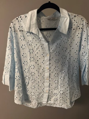 Blus - Söt ljusblå blus med broderade blommor och hålmönster. Blusen har klassisk krage, knäppning framtill och trekvartsärmar. Perfekt för en fräsch och somrig look. Tillverkad i ett lätt och luftigt tyg.