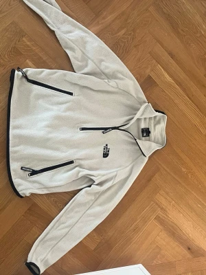  The North Face fleece - Säljer en beige fleecejacka från The North Face med svart dragkedja och logga på bröstet. Jackan har två snedställda fickor med dragkedja och hög krage. Perfekt för lager-på-lager och chill dagar.