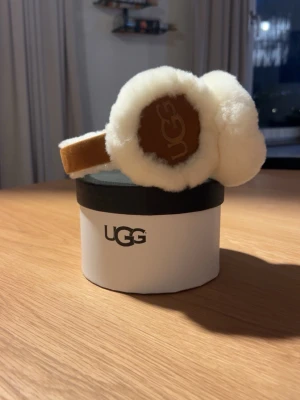 Vita öronmuffar från UGG - Mysiga öronmuffar från UGG i vit fluffig fårull med bruna detaljer och logga på sidan. Perfekta för att hålla värmen under kalla dagar och ger en trendig vinterlook.