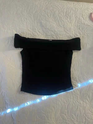 Svart offshoulder topp - Trendig svart offshoulder topp med bred kant upptill som ger en snygg och stilren look. Perfekt att matcha med både jeans och kjol för en cool outfit. Toppens passform är tight och den är ärmlös för en modern känsla.