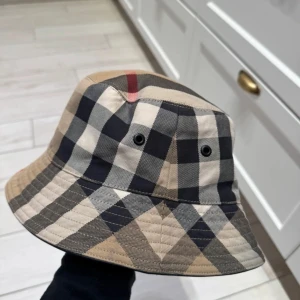 Rutig bucket hat - Snygg bucket hat från Burberry i klassiskt rutigt mönster. Hatten har en beige, svart och röd färgkombination som ger en stilren look. Perfekt för både vår och sommar. Den är i mycket bra skick och passar perfekt för en trendig outfit. Bild 3 så ser ni dock ett streck med smusts på hatten, har inte testat rengöra den då jag har tröttnat på den och inge orkat lägga ner tid och enargi. Skulle säga det är mer s än m i storleken på hatten.
