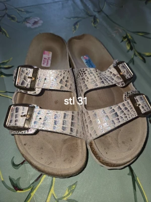 Beige sandaler med krokoprint - Säljer ett par beige sandaler från Hemma Känsla med krokodilpräglat mönster och två breda remmar med stora metallspännen. Sandalerna har en mjuk innersula och öppen tå, perfekta för en avslappnad stil.