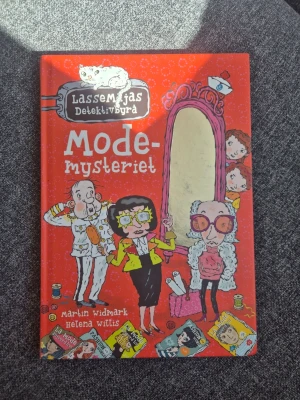 LasseMajas Detektivbyrå: Modemysteriet - En färgstark och rolig bok ur serien LasseMajas Detektivbyrå, skriven av Martin Widmark och illustrerad av Helena Willis. Omslaget är rött med tecknade figurer och spegel, och boken handlar om ett mysterium i modevärlden. Perfekt för unga deckarfans!
