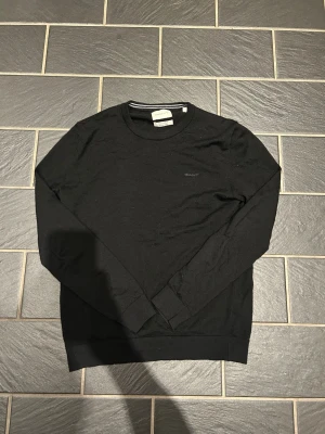 Svart tröja från Gant - Snygg svart tröja från Gant i tunn bomullskvalitet. Klassisk rund halsringning och diskret Gant-logga broderad på bröstet. Perfekt basplagg som funkar till allt och har ribbade muddar vid ärmslut och nederkant.