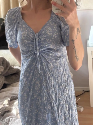 Blå blommig långklänning med puffärm - Ljusblå långklänning med små vita blommor, puffärmar och knytning framtill. Klänningen har v-ringning och är tillverkad i ett lätt, mjukt material som ger en romantisk känsla. Perfekt för sommarens alla tillfällen.