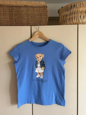 Blå Polo Ralph Lauren T-shirt med en björn på. - Blå t-shirt från Polo Ralph Lauren med björnmotiv på bröstet. Klassisk rund hals och tillverkad i mjuk bomull vilket ger materialet en bekväm  känsla.  Knappt använt tröjan. Jätte fin till sommarn. ( pris går att diskutera). 💗