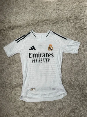 Real Madrid matchtröja Mbappé nr 9 - Säljer en vit Real Madrid matchtröja från Adidas med svarta detaljer och klubbmärke på bröstet. Tröjan har trycket 'Emirates Fly Better' fram och 'Mbappé 9' bak. Tillverkad i lätt funktionsmaterial med HEAT.RDY-teknologi och klassiska Adidas-ränder på axlarna. Storlek: XS/S