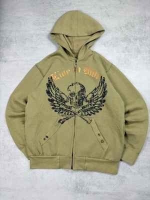 Olivgrön hoodie från Blue Brand - Säljer en olivgrön hoodie från Blue Brand med dragkedja och tryck framtill av en döskalle med vingar samt texten 'Live to Ride' i guld. Hoodien har fickor med knappdetaljer och en klassisk huva. Perfekt för dig som gillar streetwear och coola prints.