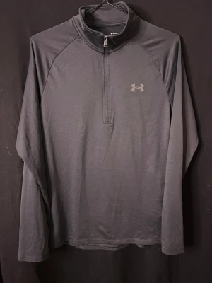 Svart långärmad träningströja UA - Snygg svart långärmad träningströja från Under Armour med halv dragkedja framtill och diskret logga på bröstet. Tillverkad i ett mjukt och stretchigt funktionsmaterial som andas, perfekt för träning eller löpning.