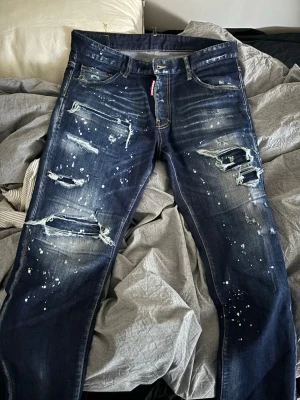 Dsquared2 blå slitna jeans herr - Säljer ett par mörkblå jeans från Dsquared2 med coola slitningar, trasiga detaljer och vita färgstänk. Säker dem då de inte passar längre. Kvitto från farfetch finns. 