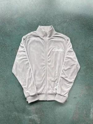 Grå velour tracksuit från Crtz corteiz - Snygg grå ziptröja i mjuk velour med matchande byxor från Crtz med vit broderad logga på bröstet och stor logga på ryggen. Tröjan har hög krage, långa ärmar och muddar vid ärmslut och midja. Perfekt för en chill och trendig look. PRIS KAN DISKUTERAS!!