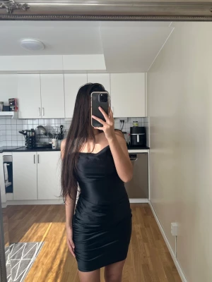 Svart satin kortklänning med smala band - Snygg svart kort klänning i satin med smala axelband och figurnära passform. Klänningen har en lätt glansig yta och enkel, stilren design som ger en elegant look. Perfekt för dig som gillar minimalistisk stil och vill sticka ut.