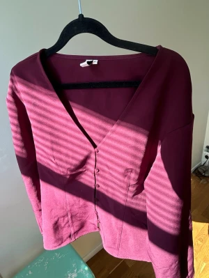 Vinröd kofta med knappar från Nelly - Snygg vinröd kofta med v-ringning och knäppning framtill. Modellen har långa ärmar och är gjord i ett mjukt, stretchigt material. Perfekt att slänga över en topp för en chill look.🥂