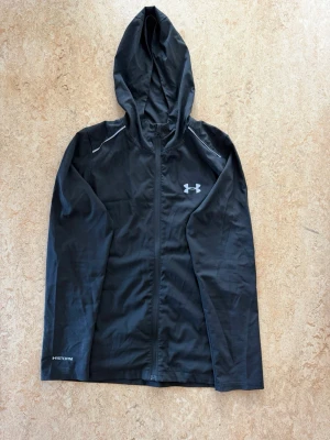 Svart vindjacka från Under Armour - Snygg svart vindjacka från Under Armour med huva och hel dragkedja. Jackan har vita detaljer vid axlarna och logga på bröstet samt texten 'STORM' på ärmen. Tillverkad i lätt och vattenavvisande material, perfekt för blåsiga dagar.