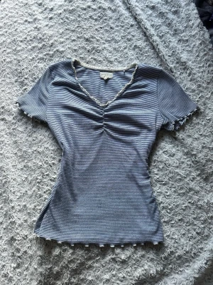 Randig topp från Holly & Whyte - Säljer en söt topp från Holly & Whyte med blå och vita ränder. Toppen har korta ärmar, v-ringning med rynkdetalj framtill och vågiga kanter. Den är lite insydd som gör att den ger en mer figursydd fin look. Står att den är en stolek L men passar mer än S-M