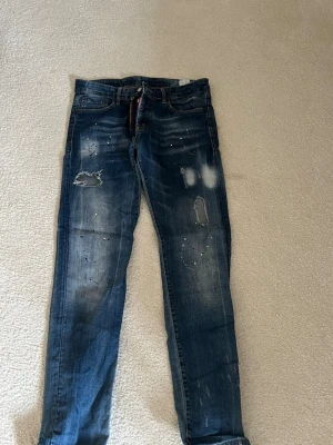 Blå distressed jeans med färgstänk - Säljer ett par blå jeans med slitningar, hål och coola färgstänk. Jeansen har klassisk femficksmodell, dragkedja och knapp i midjan. Materialet är denim med tvättad look och detaljer som ger en trendig och avslappnad stil.