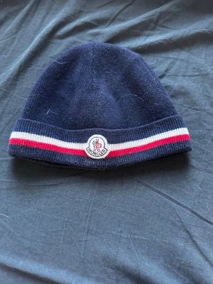 Mörkblå Moncler mössa med ränder - Snygg mörkblå mössa från Moncler med klassiska röda och vita ränder runt kanten. Mössan har Moncler-loggan framtill och är stickad i mjukt material som känns lyxigt och bekvämt. Perfekt accessoar för kalla dagar och stilrena outfits. Den är liten i storlek