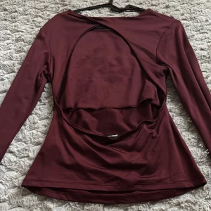 Vinröd långärmad topp med öppen rygg - Säljer en vinröd långärmad topp med unik öppen rygg-detalj. Toppen har en tight passform och är tillverkad i ett stretchigt syntetmaterial som ger en snygg siluett. Perfekt för dig som vill sticka ut med en cool och trendig look.