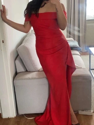 Röd offshoulder långklänning - Fin röd långklänning med offshoulder-design och figurnära passform. Klänningen har en snygg slits framtill och draperade detaljer längs midjan och höften.