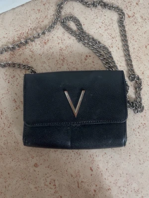 Svart axelväska med kedjerem - Snygg svart axelväska i konstläder med silverfärgad kedjerem och ett V-format metallspänne på framsidan. Väskan har en stilren och kompakt design som passar perfekt till många outfits. Perfekt för dig som vill ha en trendig och praktisk väska.