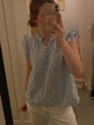 Ljusblå volangblus med knappar - Söt ljusblå blus med volangdetaljer vid axlarna och korta ärmar. Blusen har en liten ståkrage och knappar framtill. Tillverkad i ett tunt, luftigt tyg som passar perfekt till sommaren. Enkel och stilren design.