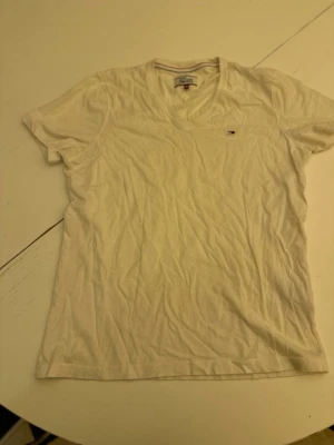 Vit v-ringad t-shirt Tommy Hilfiger - Klassisk vit t-shirt från Tommy Hilfiger Denim med v-ringning och liten broderad logga på bröstet. Tillverkad i mjuk bomull och har en normal passform. Perfekt basplagg för alla tillfällen.