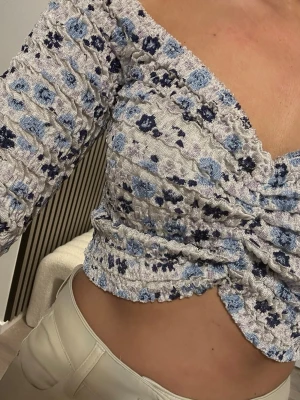 Blommig offshoulder croppad topp - Trendig offshoulder topp med croppad passform och v-ringning. Toppen har ett blått och mörkblått blommigt mönster på ljus botten, samt rynkade detaljer framtill. Perfekt för dig som vill sticka ut med en söt och modern look.
