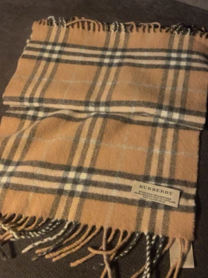 Beige rutig halsduk från Burberry - Klassisk beige halsduk från Burberry med ikoniskt rutmönster i svart, vitt och rött. Halsduken har fransar i ändarna och är tillverkad i mjuk ull. Perfekt accessoar för att lyfta din outfit med en tidlös touch.