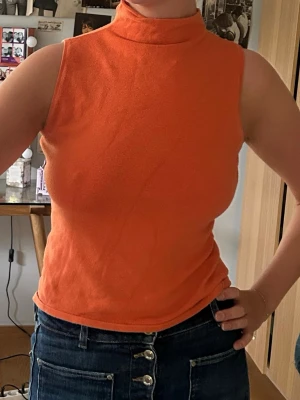 Orange ärmlös polotopp - Säljer en snygg orange polotopp i mjukt material. Toppen är ärmlös och har en hög krage som ger en trendig look. Perfekt att styla med jeans eller kjol för en cool vibe. Köpt på en vintage butik i Stockholm 