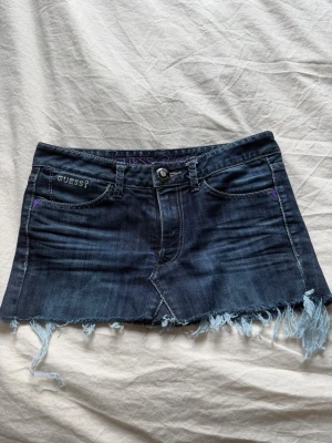 Mörkblå jeanskjol från Guess - Snygg mörkblå jeanskjol från Guess med fransig nederkant och klassiska jeansdetaljer. Kjolen har knapp och dragkedja framtill samt fickor både fram och bak. Perfekt för dig som gillar en edgy och trendig stil.