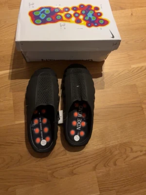  Säljer ett par Svarta Nike mind tofflor - Säljer ett par svarta Nike tofflor med ventilerande mesh på ovansidan och coola orange detaljer på sulan. Insidan har en färgglad design med prickar och Nike Mind-tryck. Perfekta för chill dagar hemma eller efter träningen.
