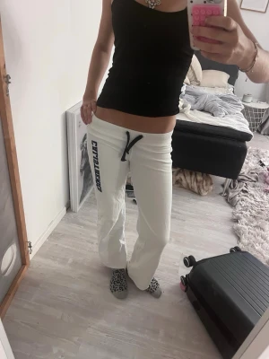 Vita mjukisbyxor med tryck - Säljer ett par vita mjukisbyxor med svart snörning i midjan och ett stort tryck längs benet. Byxorna har rak passform och är perfekta för chill dagar hemma eller när du vill vara bekväm. Materialet ser ut att vara mjukt och stretchigt.