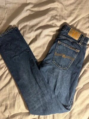 Nudie Jeans - Säljer ett par klassiska blå jeans från Nudie Jeans Gritty Jackson med raka ben och femficksmodell. Jeansen har snygga kontrastsömmar och en ikonisk bakficka med broderad våg. Tillverkade i slitstark denim, perfekta för en avslappnad stil.