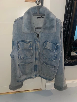 Ljusblå jeansjacka med fuskpäls - Snygg ljusblå jeansjacka med fyra stora fickor framtill och mjuk fuskpäls på kragen. Jackan har dragkedja och uppvikta ärmslut för en avslappnad look. Perfekt för dig som vill ha en trendig och bekväm jacka med extra detaljer.