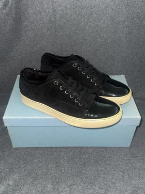 Svarta Lanvin sneakers med mocka  - Snygga svarta Lavins i mocka med lackad tå och vit sula. Skorna har klassisk låg profil, svarta skosnören och metallöljetter. Perfekta för dig som gillar stilrena detaljer och en clean look.