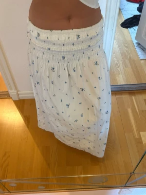 Vit sommarklänning med blå blommor - Söt axelbandslös klänning i vitt med små blå blommor och smock upptill. Klänningen är vadlång och har en luftig passform, perfekt för varma dagar. Materialet ser ut att vara bomull eller en bomullsblandning. Romantisk och enkel stil.