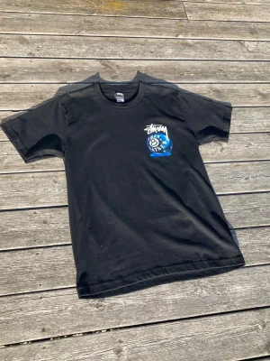 Svart Stüssy t-shirt med tryck - Svart t-shirt från Stüssy med ett blått och vitt tryck på bröstet som föreställer en magisk 8-ball och texten 'Born Raised'. Klassisk passform och rund halsringning, perfekt för en streetwear-look.
