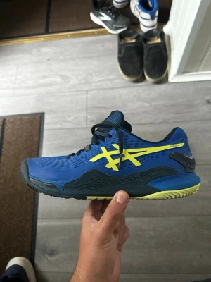 Blåa Asics Padelskor med gula detaljer - Säljer ett par blåa Asics Padelskor med tydliga gula detaljer och svart sula. Skorna har låg profil, meshmaterial för bra ventilation och snörning framtill. Perfekta för dig som vill ha snygga och sportiga skor till padel 