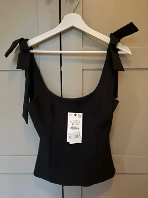 Svart linnetopp med rosetter - Stilren svart linnetopp med breda axelband som knyts i snygga rosetter på axlarna. Toppen har en figurnära passform och är perfekt för att skapa en chic look. Tillverkad i ett mjukt och stretchigt material som känns bekvämt hela dagen.
