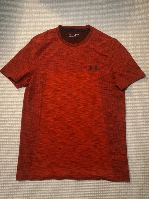 Röd träningst-shirt från Under Armour - Snygg röd t-shirt från Under Armour med diskret mönster och svart logga på bröstet. Perfekt för träning med sitt lätta och ventilerande material. Klassisk passform och rund halsringning.