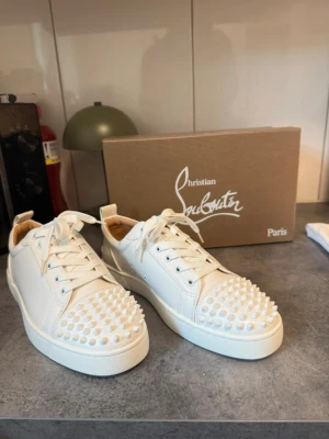Vita sneakers med nitar från Louboutin - Säljer ett par vita sneakers från Christian Louboutin med coola nitar över tån. Skorna har snörning och är tillverkade i skinn med en chunky sula. Perfekta för dig som vill sticka ut med en edgy och lyxig stil. Helt nya oanvända 