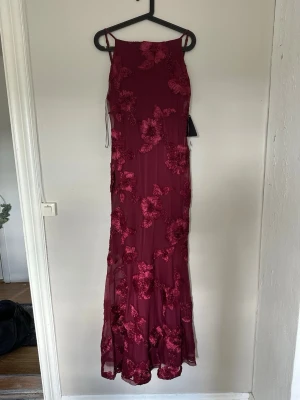 Vinröd långklänning med blommor - Elegant vinröd långklänning med tunna axelband och blommiga applikationer i samma ton. Klänningen har en figurnära passform och är tillverkad i ett lätt, transparent material med blommiga detaljer som ger en lyxig känsla. Slutsåld!