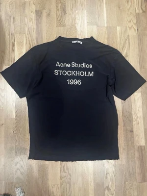 Svart Acne Studios t-shirt - Svart t-shirt från Acne Studios med tryck 'Acne Studios STOCKHOLM 1996' på bröstet. Klassisk rak passform och rund hals. Tillverkad i mjukt bomullstyg som känns skönt mot huden. Perfekt för en clean och stilren look.